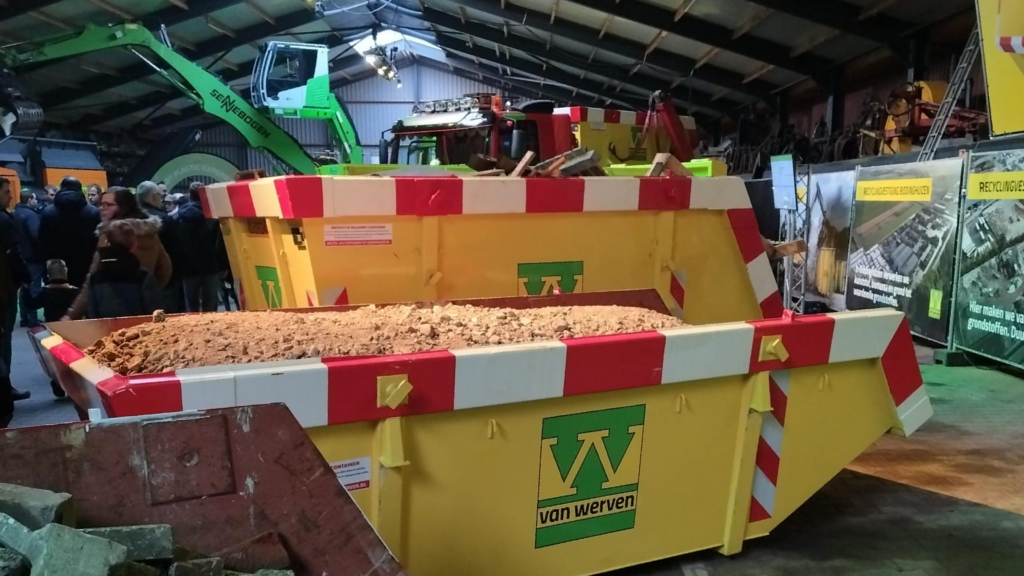 Een gele afvalcontainer met aarde in een grote hal vol mensen.