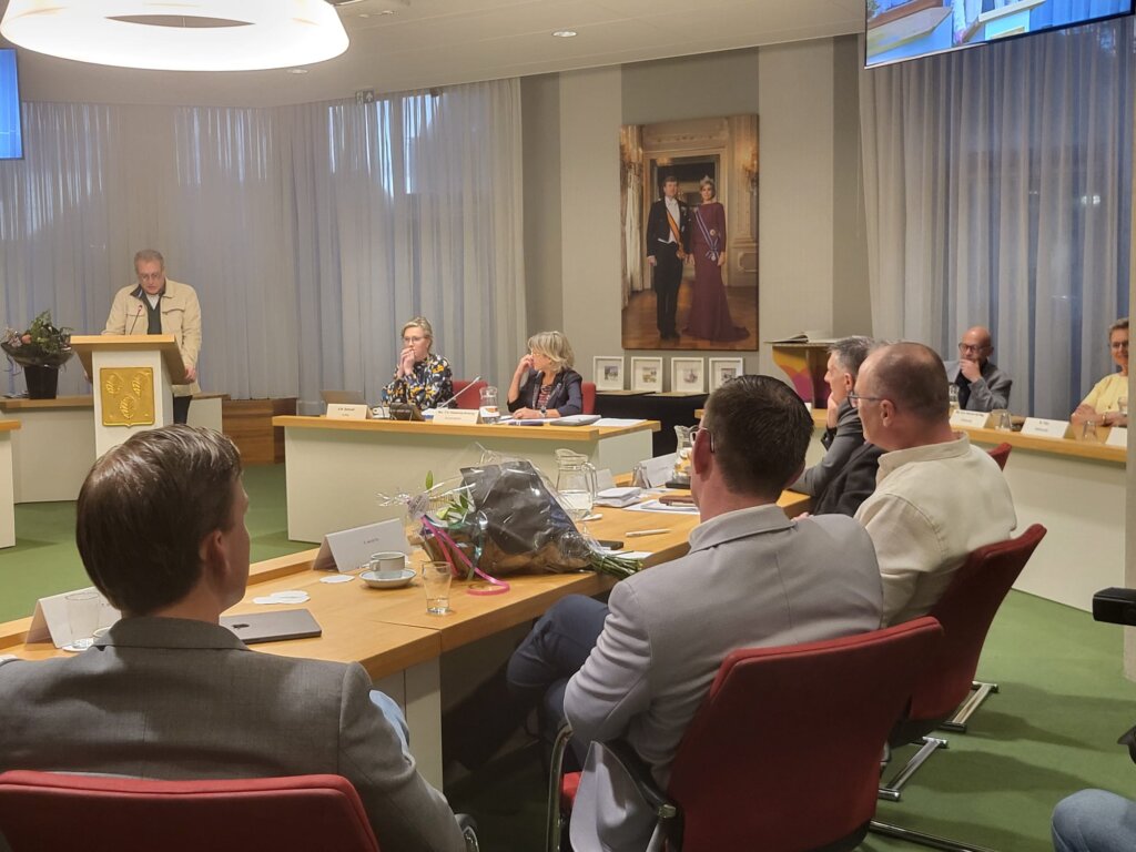 Een formele vergadering met sprekers en publiek in een zaaltje.