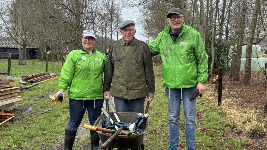 Drie mensen staan buiten in groene jassen, met een kruiwagen op de achtergrond.